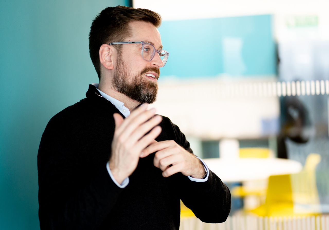 Man with a beard and glasses speaking and gesturing with his hands in a modern office setting