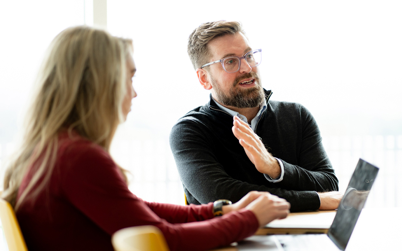 Man with glasses sat at an office desk with a female colleague explaining something to the room